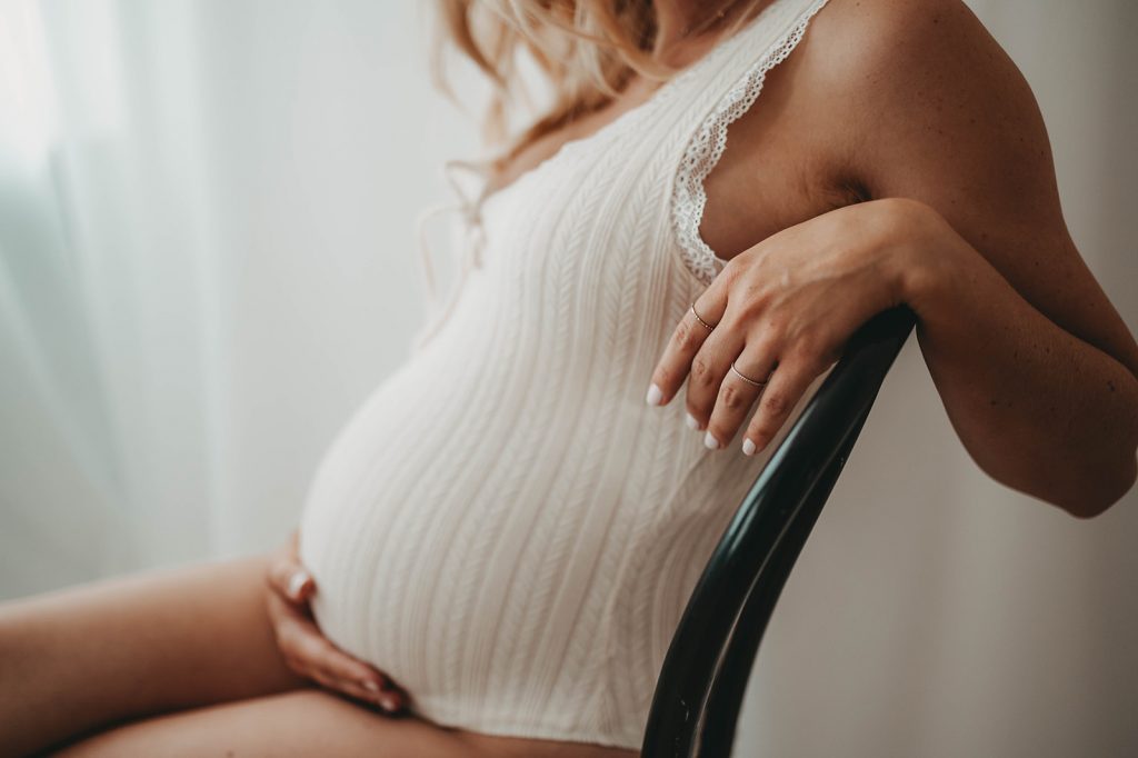 Schwangere Frau sitzt in einem hellen Body auf einem schwarzen Stuhl und hält ihren Babybauch