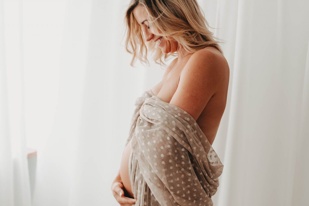 Nackte schwangere Frau mit einem braungrauen Tuch bedeckt lächelt und schaut auf ihren nackten Babybauch
